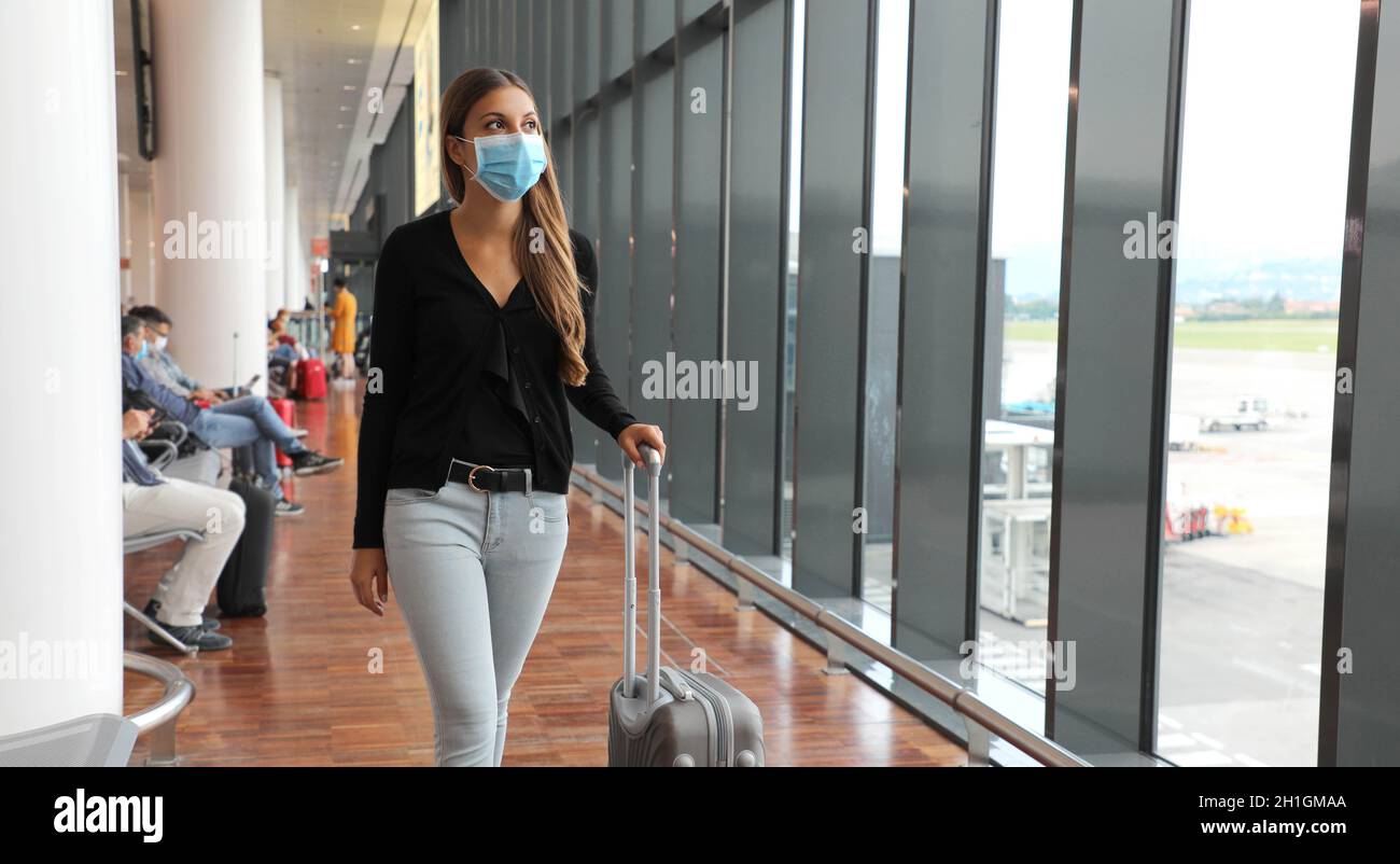Young business woman with surgical mask and travel trolley luggage walking inside the airport. Female executive with suitcase on business trip during Stock Photo