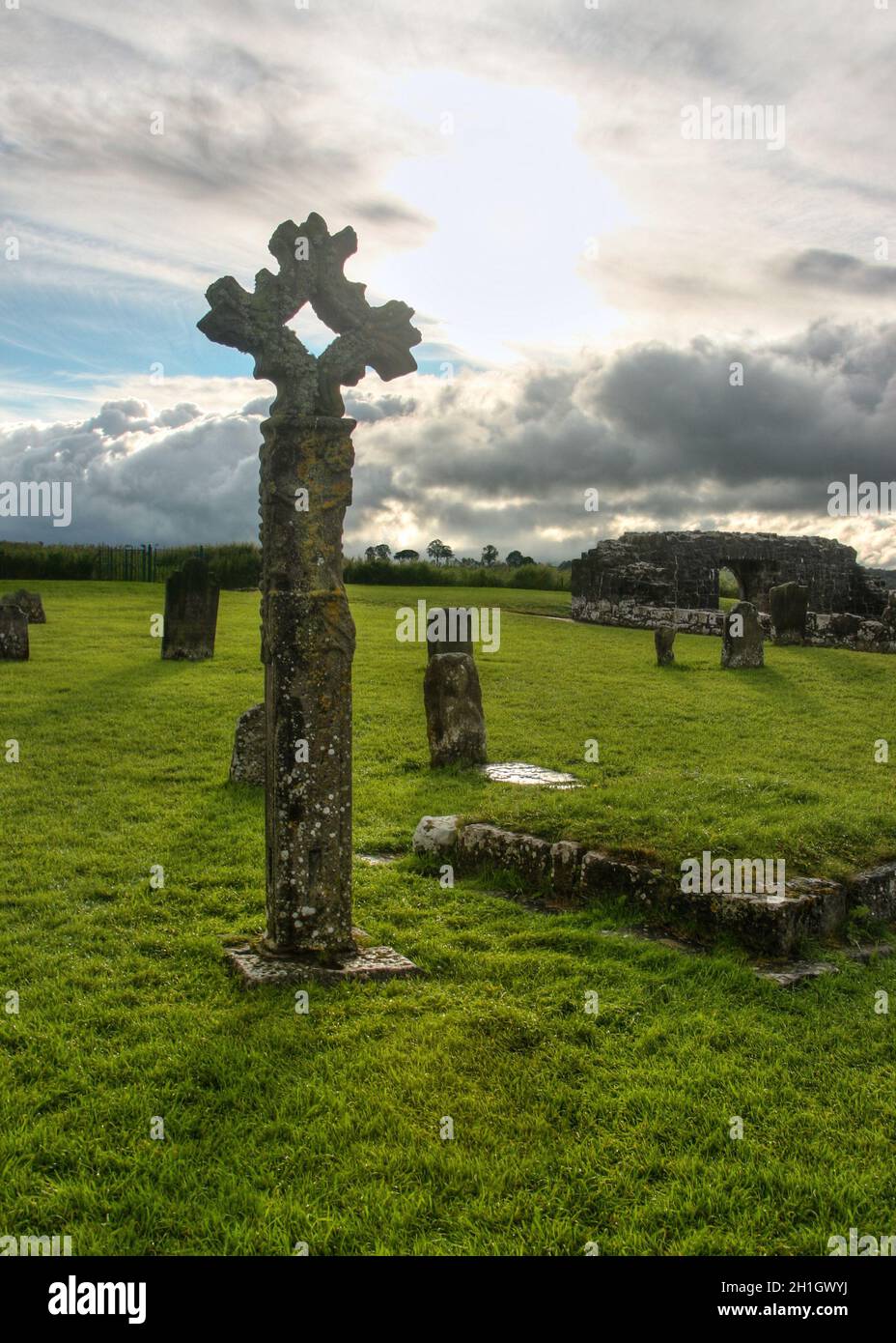 Old celtic stone cross on cemetery full of green grass with some ruins and sun creating backlight through clouds in background. Devenish Island, Ennis Stock Photo