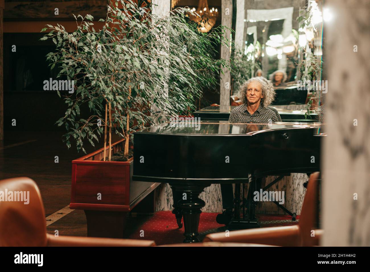 Photo of 70-year-old man plays grand piano in lobby of hotel view from afar, gray curly hair. Stock Photo