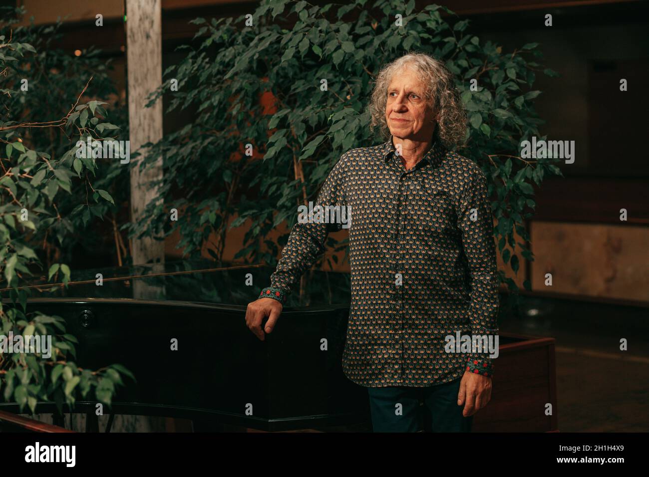 portrait of 65-year-old man at piano musical instrument in lobby of hotel among green trees gray curly hair, charismatic appearance Stock Photo