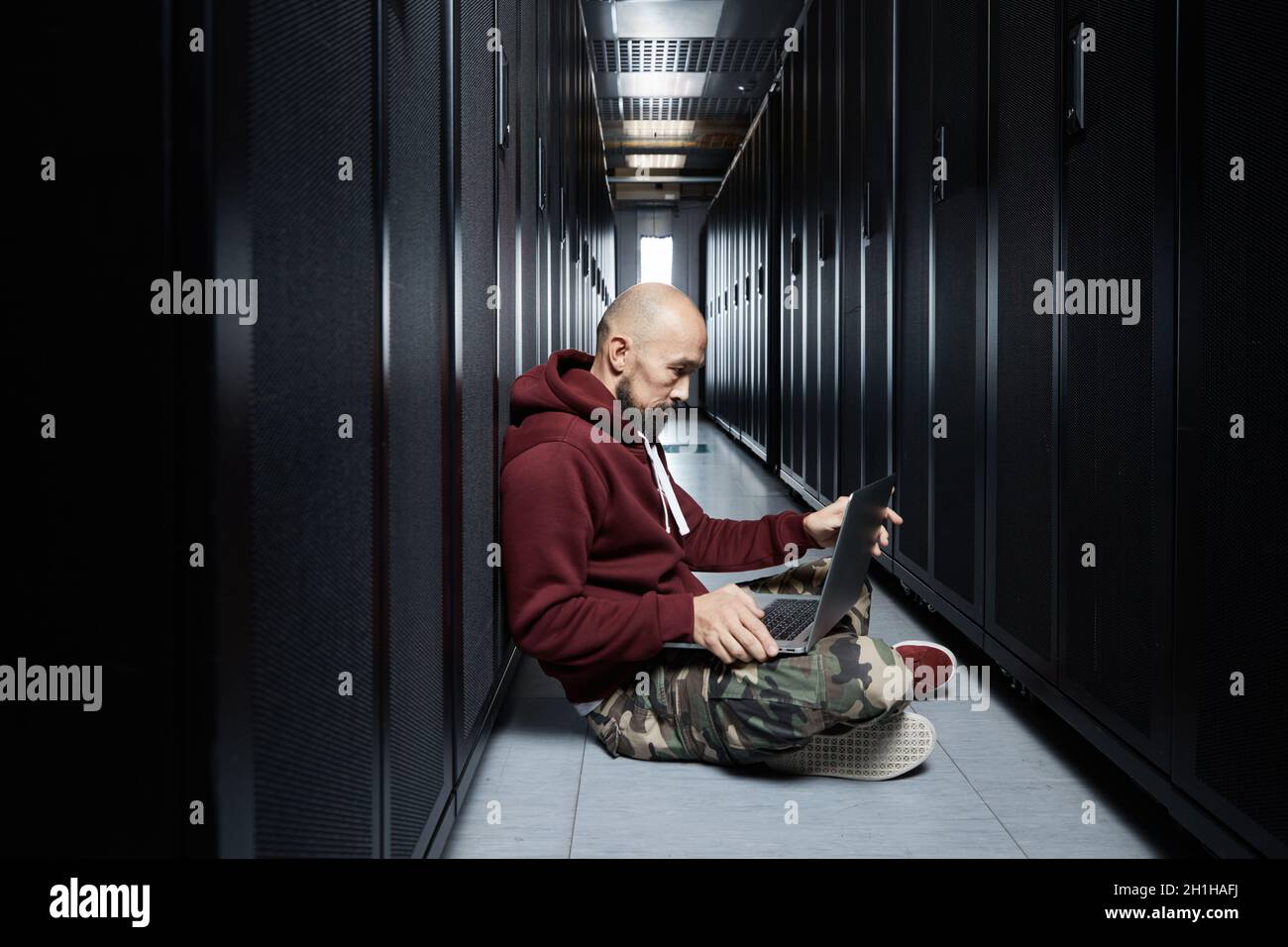 The hacker with a bald head and a beard sits with the computer among servers, the burglar, Storage of data, cloudy services, system breaking, attack Stock Photo