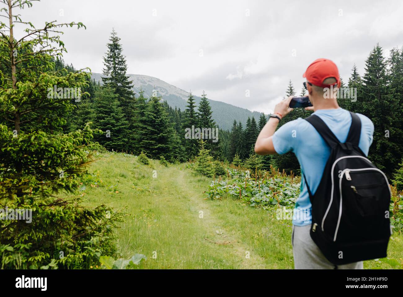 A young man with a backpack takes pictures of the mountain top on his phone and enjoys a beautiful view of the mountains. Stock Photo