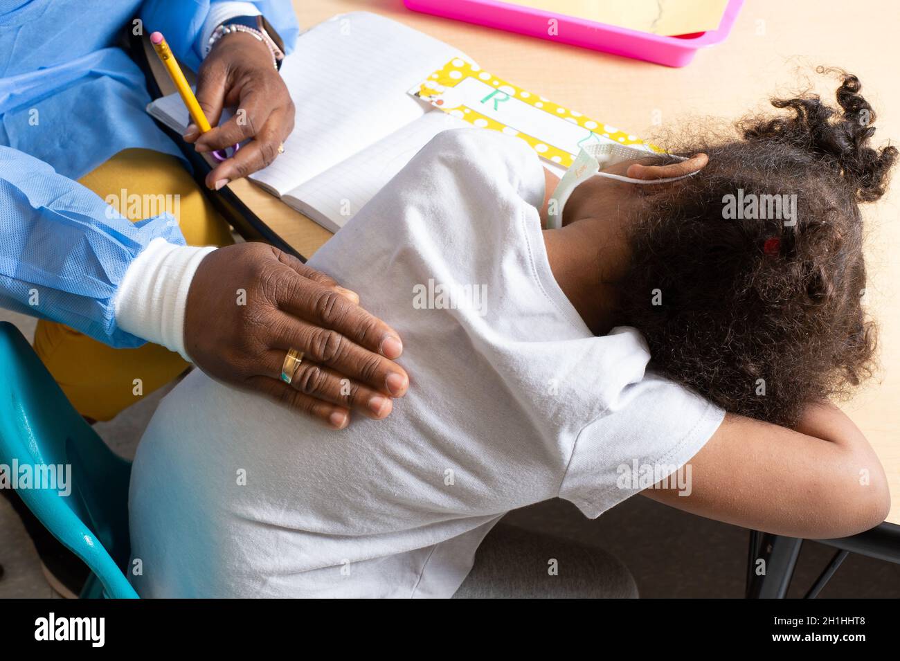 Education Preschool 3-4 year olds girl laying head on table, sad or tired at start of school day, teacher patting her on the back, wearing face mask Stock Photo