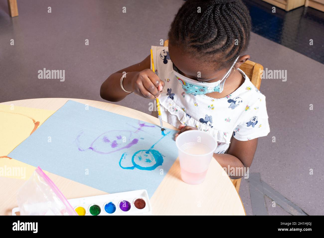 Education Preschool 4-5 year olds girl sitting at table painting with water colors, using right hand, wearing face mask Stock Photo