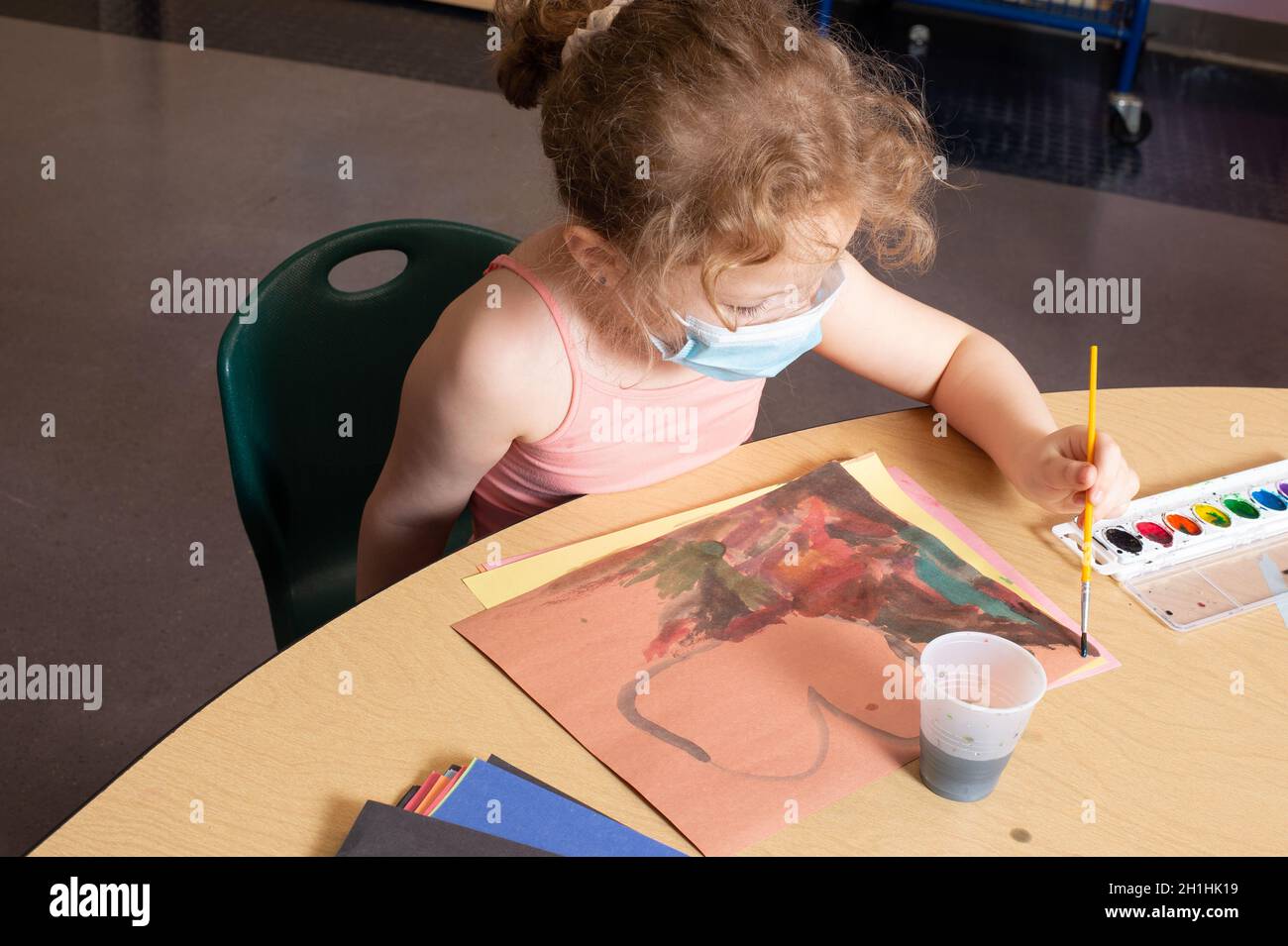 Education Preschool 4-5 year olds girl sitting at table painting with water colors, using left hand, wearing face mask Stock Photo