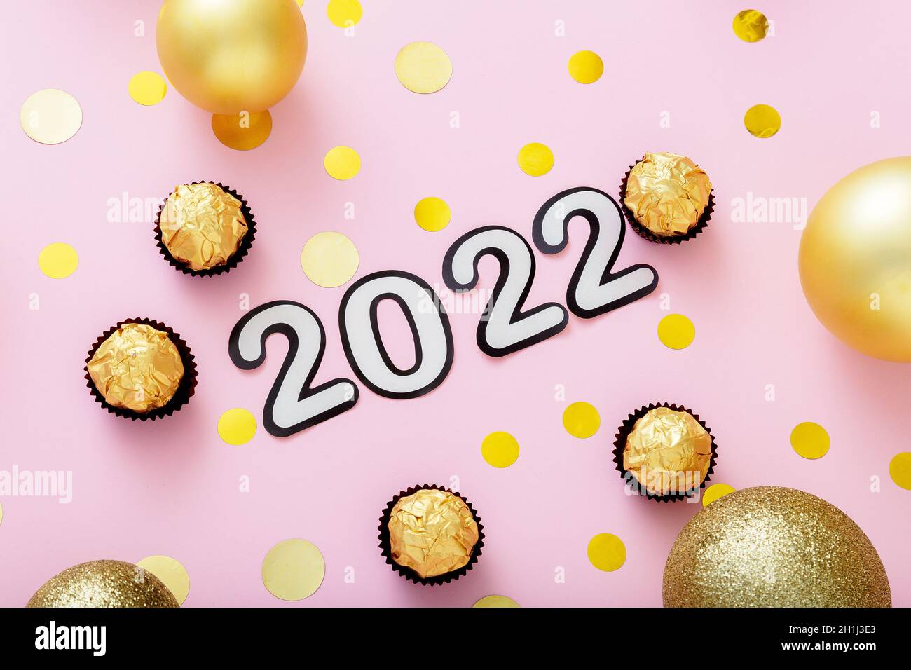2022 numeral lettering in Happy New Year style. 2022 text with Christmas decorations candies Sweets gold confetti on pink color background. Stock Photo