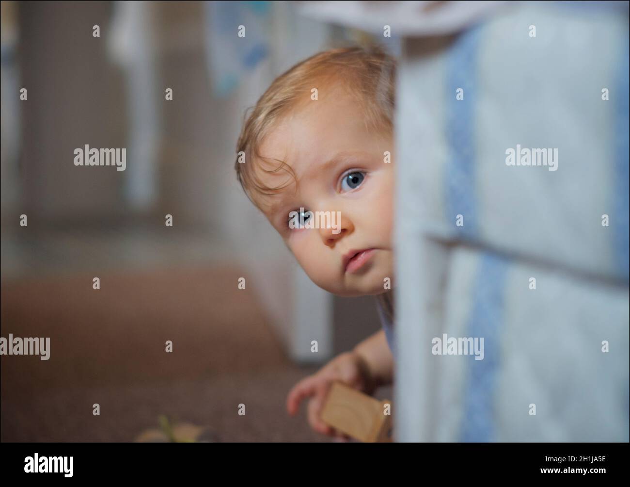 Portrait of an inquisitive baby, peering out from behing the bed, with toy in hand. Stock Photo