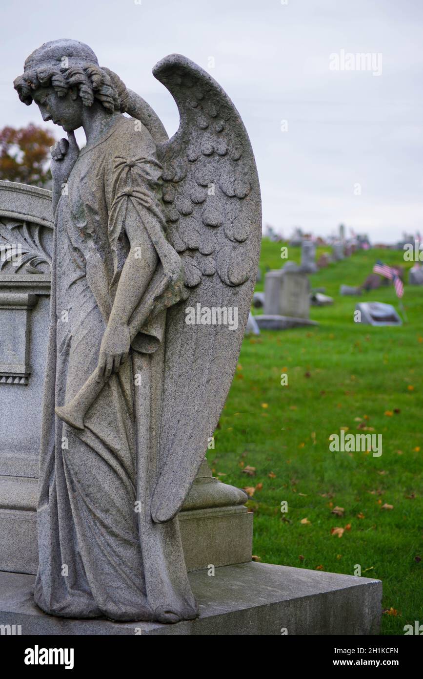 Sad angel with big wings and a trumpet stands off center with head bowed by a grave. Natural light with copy space, defocused background Stock Photo