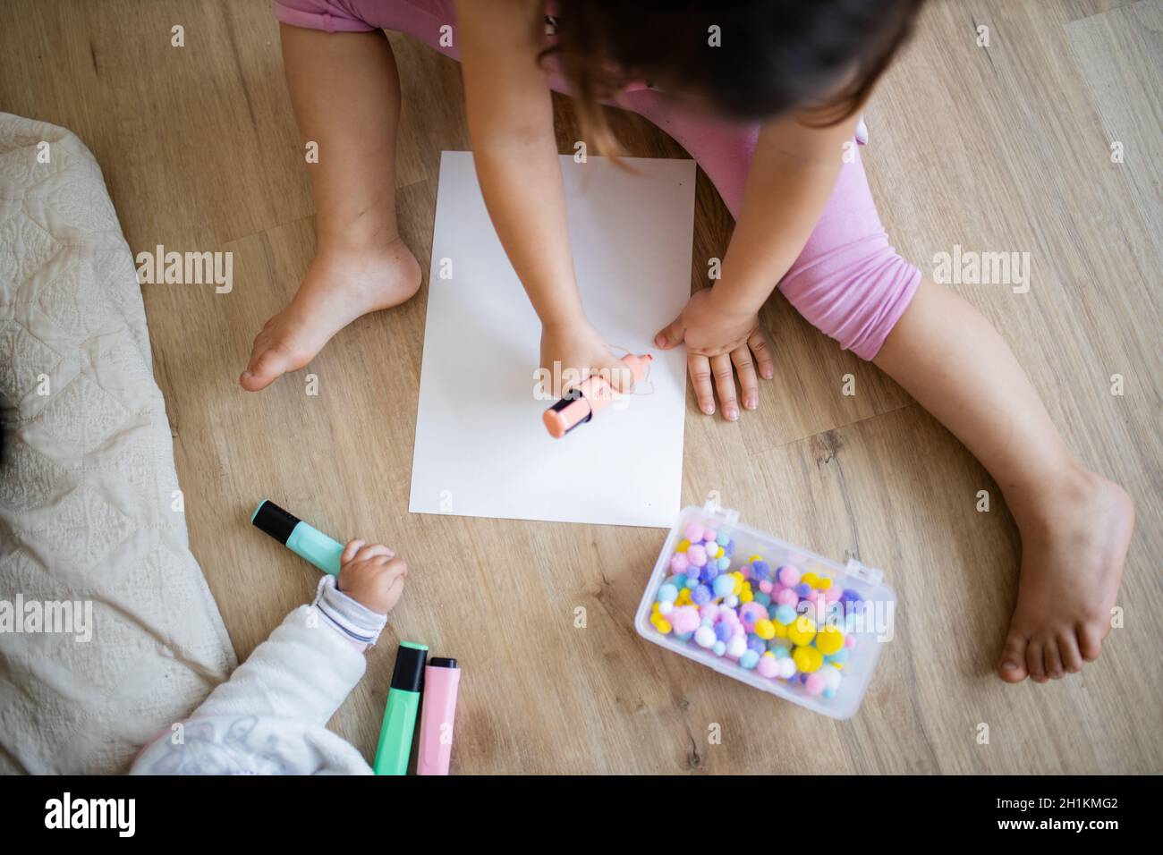 Landscape picture of a little girl in pink clothing sitting on a wooden floor and doing sums on a paper sheet with a pink textmarker and colorful cott Stock Photo