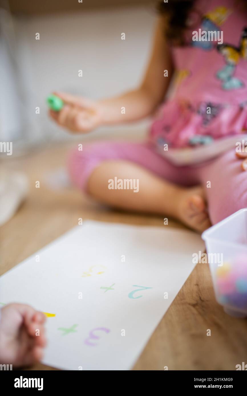 Portrait picture of a little girl in a pink dress sitting on the wooden floor and doing sums on a paper sheet with a green textmarker, alongside a pla Stock Photo