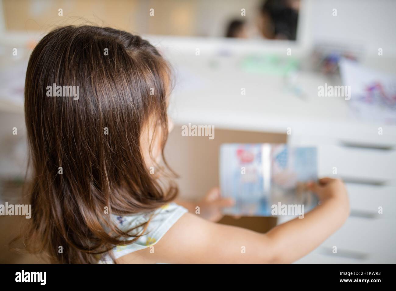 Picture over the shoulder of a little brunette girl looking at the colorful seals of an American passport, and with a blurry mirror and a desk full of Stock Photo