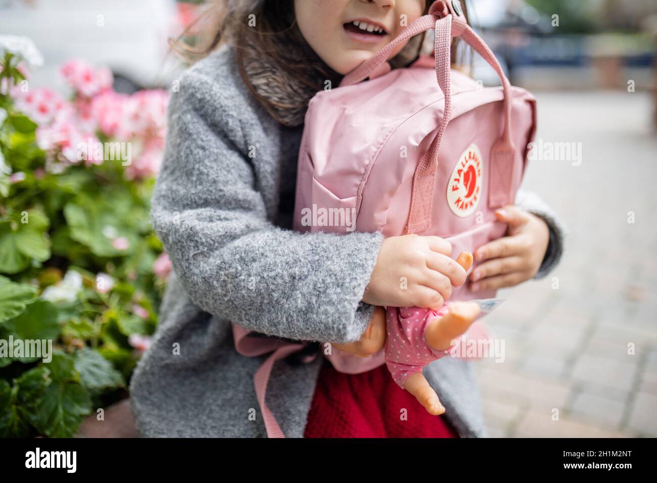 London, UK - November 4, 2020: Happy little girl with flowers behind her hugging pink Fjallraven backpack and small doll. Young child holding pink ruc Stock Photo