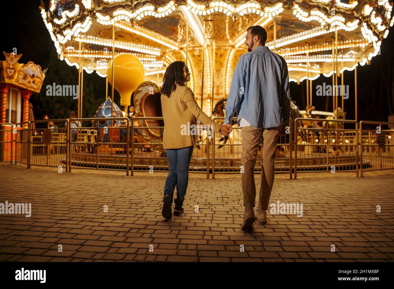 Love couple at the carousel in night amusement park. Man and woman relax outdoors, roundabout attraction with lights on background. Family leisures in Stock Photo