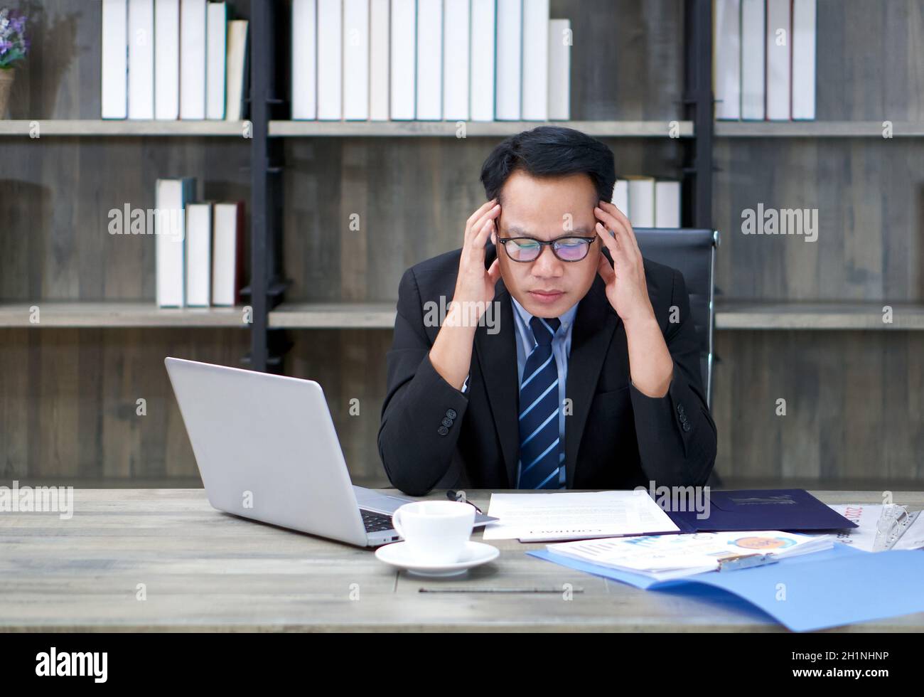 Sad Asian businessman holds his temple with both hands, sitting in the office. The concept of unemployed, sadness, depressed and human problems. Stock Photo