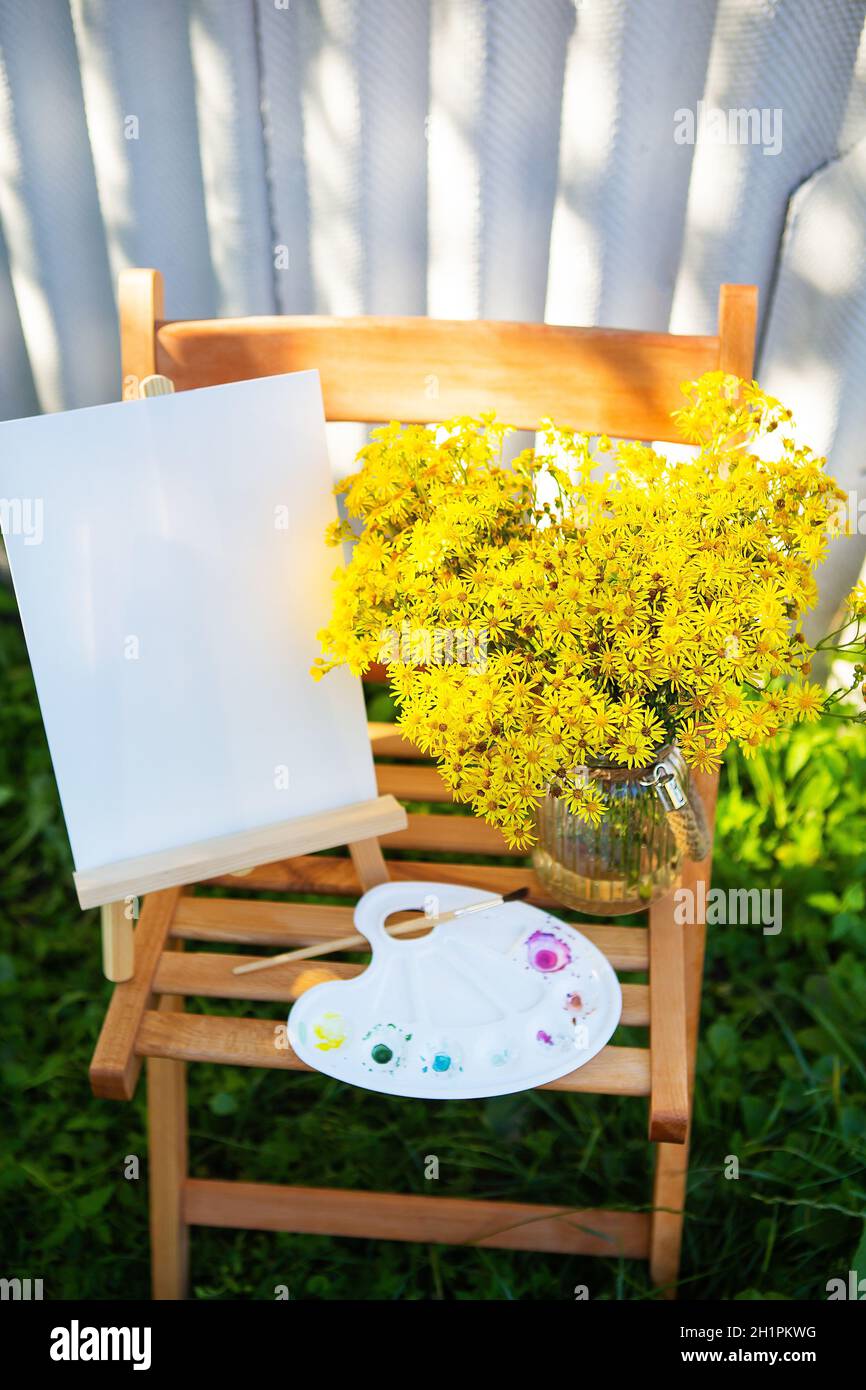 A palette of watercolor multicolored paints stands on a wooden chair and a bouquet of yellow flowers, creative hobby, drawing on the street. Sunny day Stock Photo