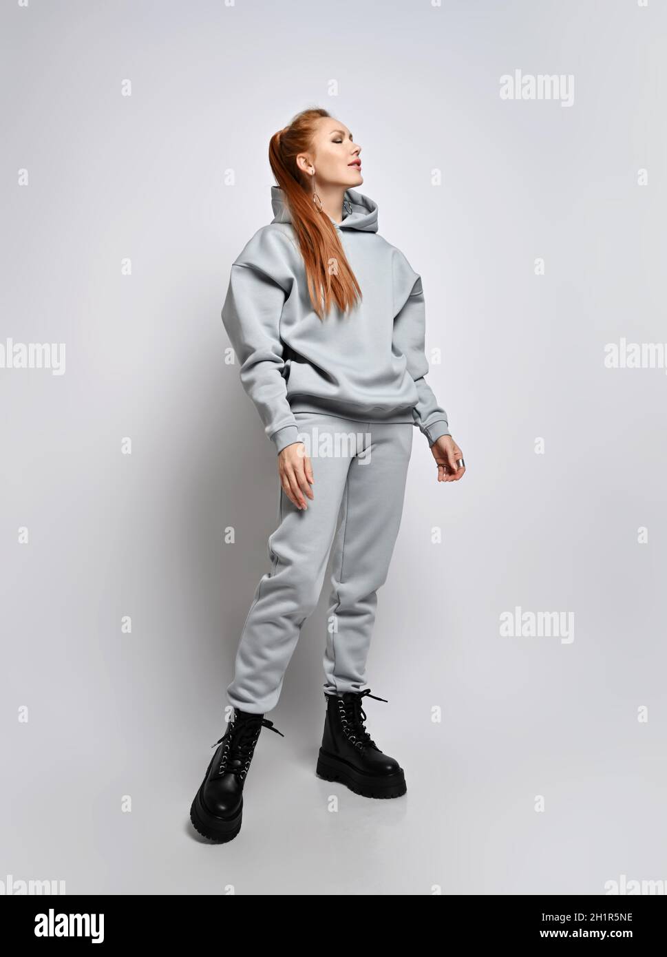 Rich slim red-haired woman in trendy grey sportswear hoodie, pants and black shoes stands sideways with eyes closed Stock Photo