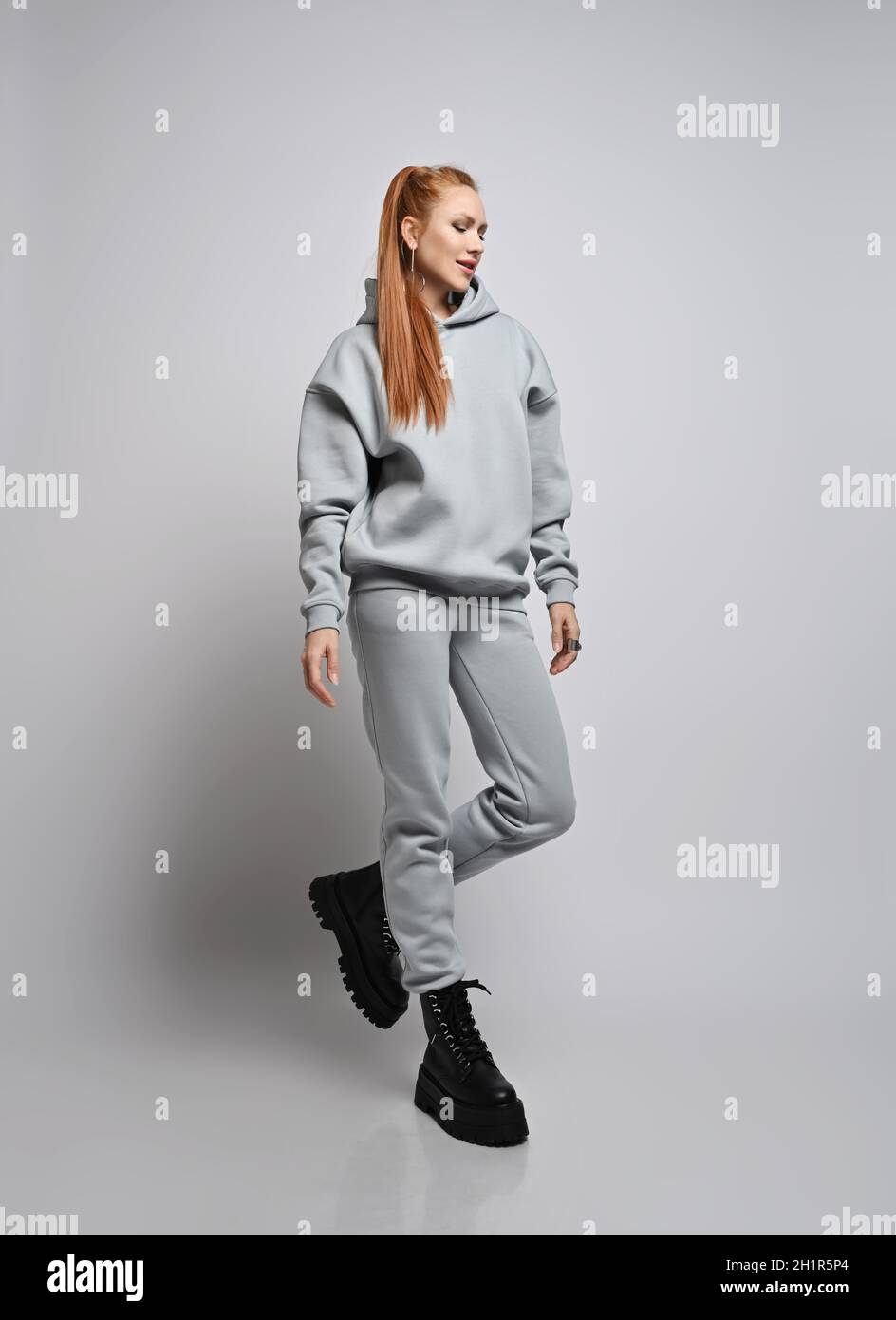 Happy rich slim red-haired woman in trendy grey sportswear hoodie, pants and black shoes walks, looking down Stock Photo