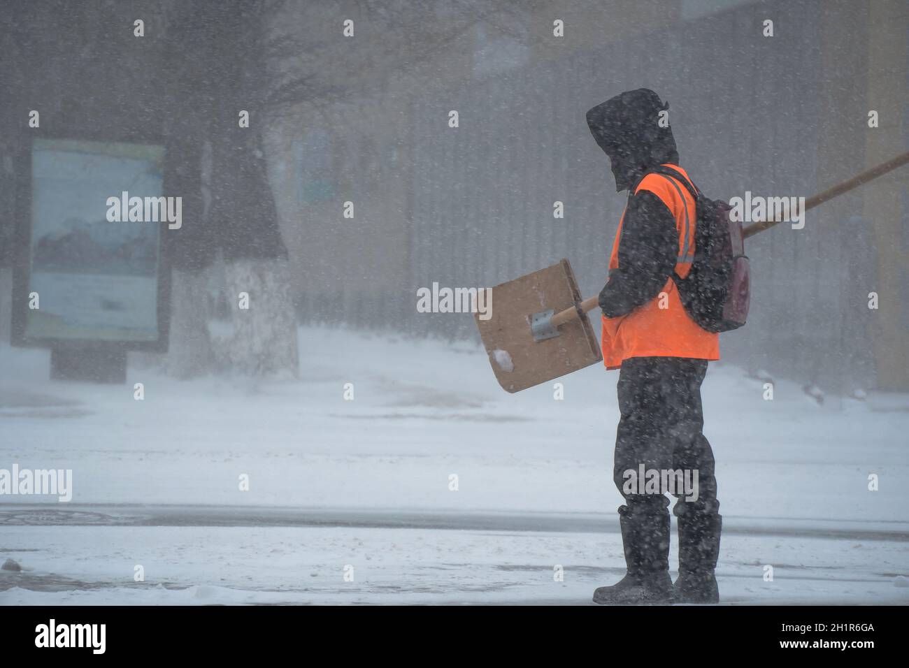 A working man of the municipal service with a snow shovel walks along the road in a storm, blizzard or snowfall in winter in bad weather in the city.E Stock Photo