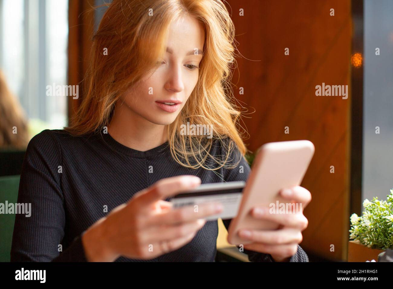 Red haired young woman making card payment through mobile phone to pay bills at a cafe. Close up. Stock Photo