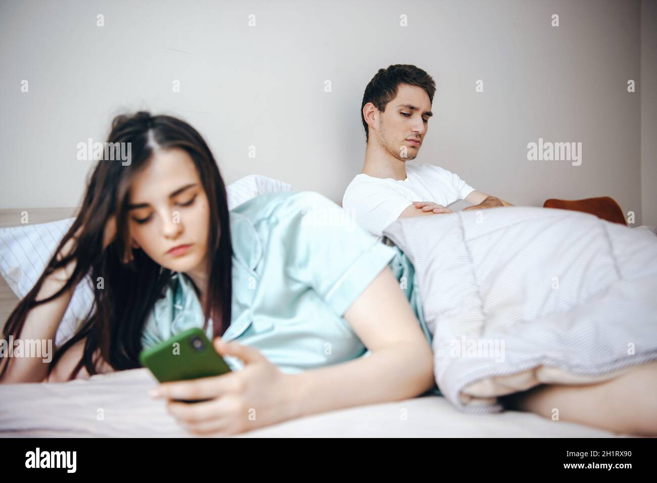 Couple sitting in bed arguing and now they are upset with each other, the woman having turned her back on the man writing messages on the phone Stock Photo