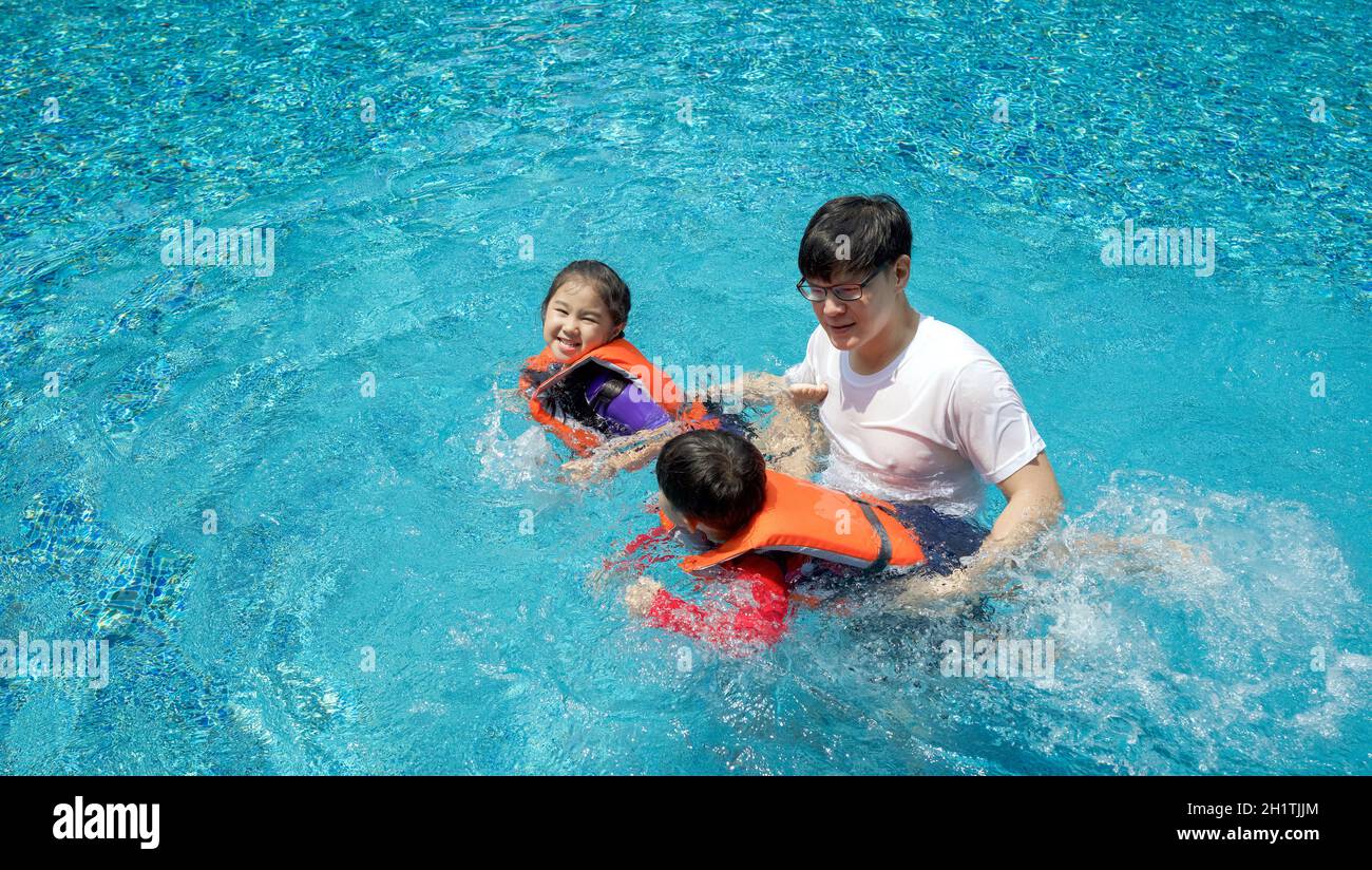 An Asian father teaches his son and daughter to swim in the pool. The older sister and younger brother wearing orange life jacket. Stock Photo