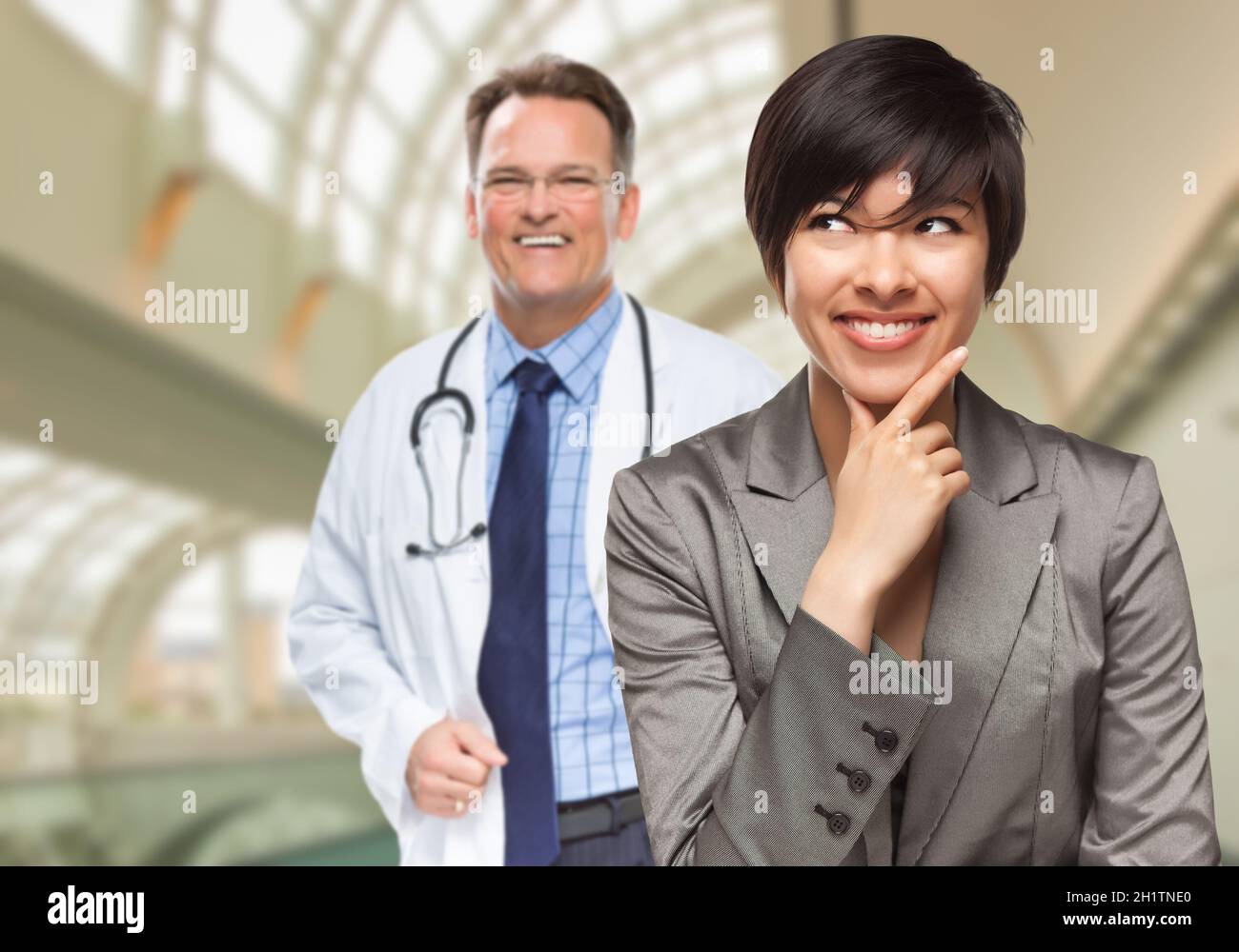 Happy Mixed Race Woman Looking To The Side As Male Doctor Stands Behind Her Inside Hospital. Stock Photo