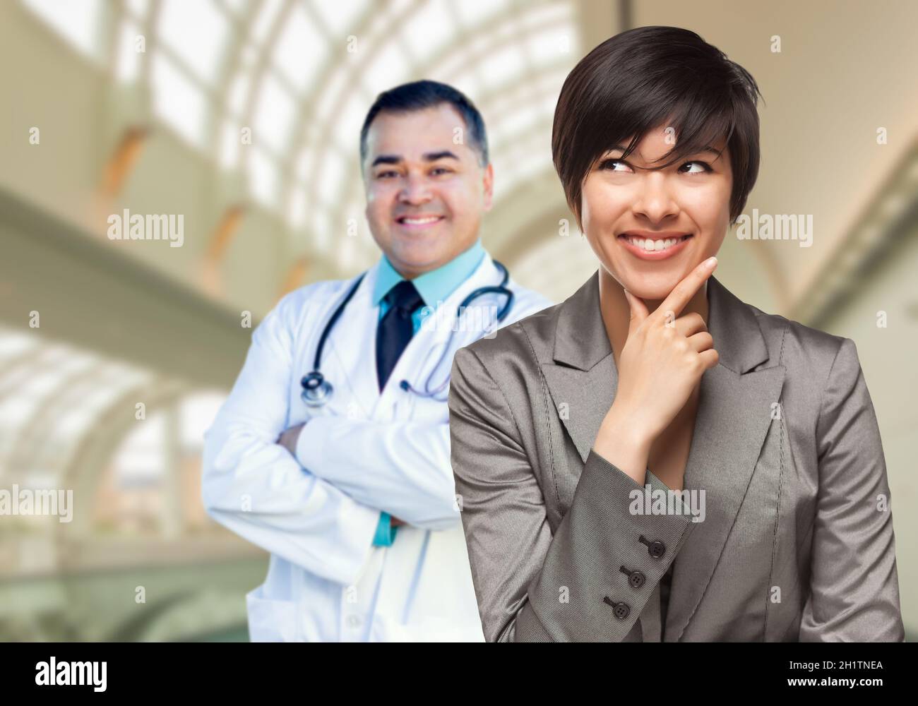 Happy Mixed Race Woman Looking To The Side As Hispanic Male Doctor Stands Behind Her Inside Hospital. Stock Photo