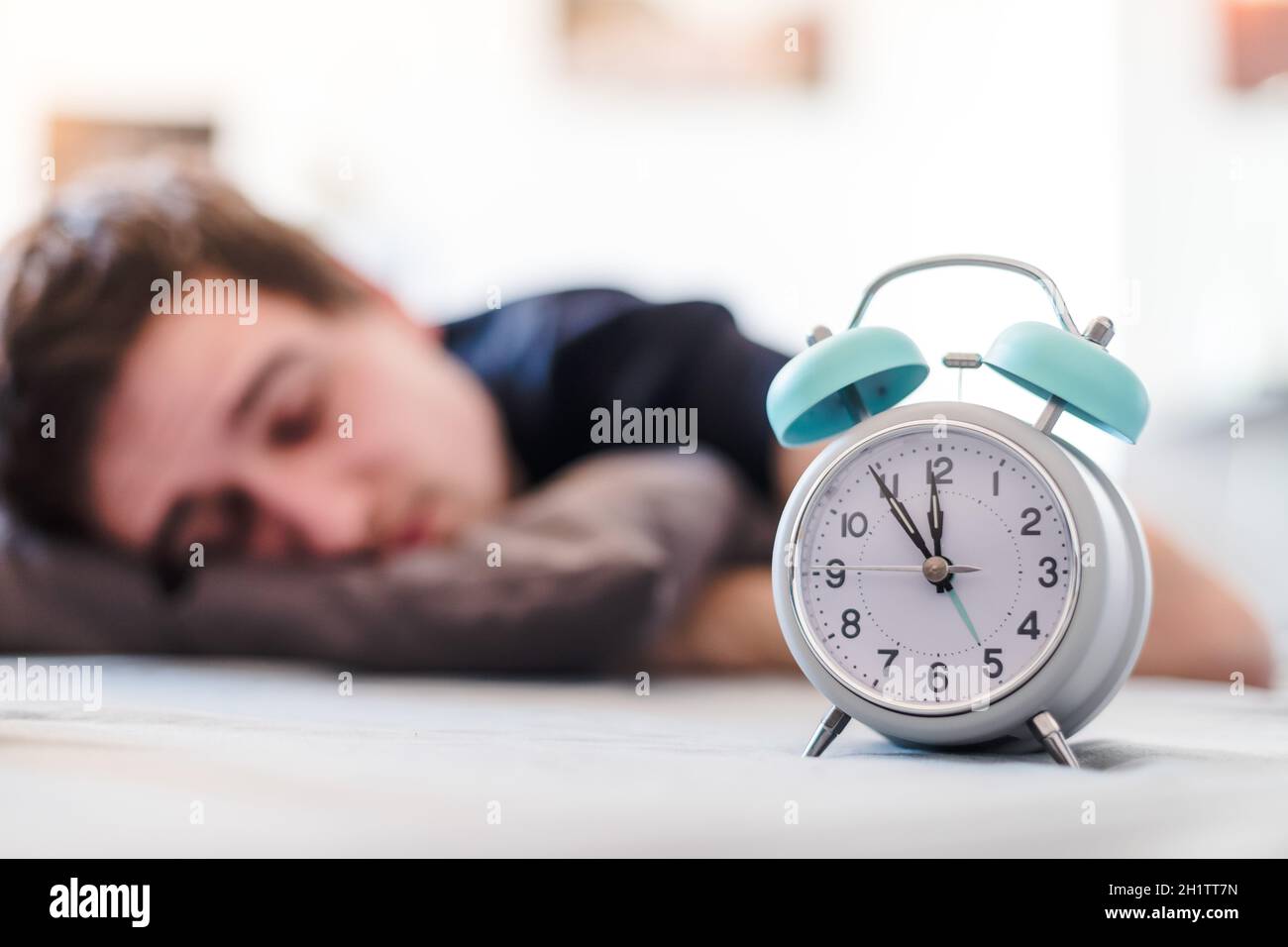 White alarm clock in the morning. Young man sleeps in the background. Stock Photo
