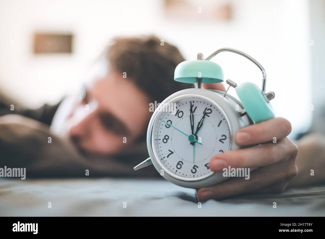 White alarm clock in the morning. Young man sleeps in the background. Stock Photo