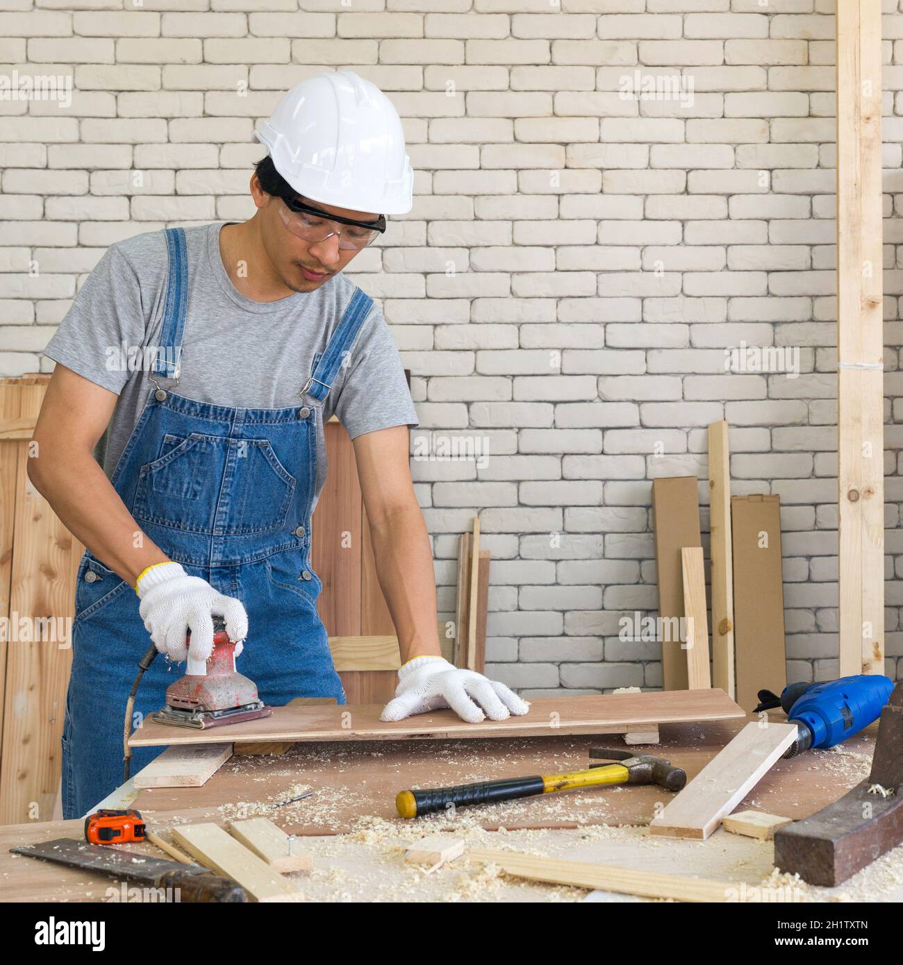 Asian carpenter with a hard hat and dustproof glasses use a sander machine to smooth plywood surface by abrasion with sandpaper. Morning work atmosphe Stock Photo