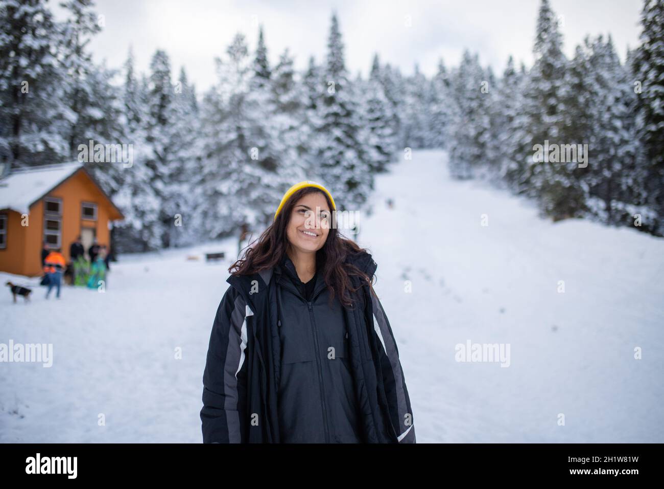 Happy woman standing with a beautiful snowy forest and small cabin as background. Smiling woman surrounded by snowy pine trees. Adventurous winter hol Stock Photo