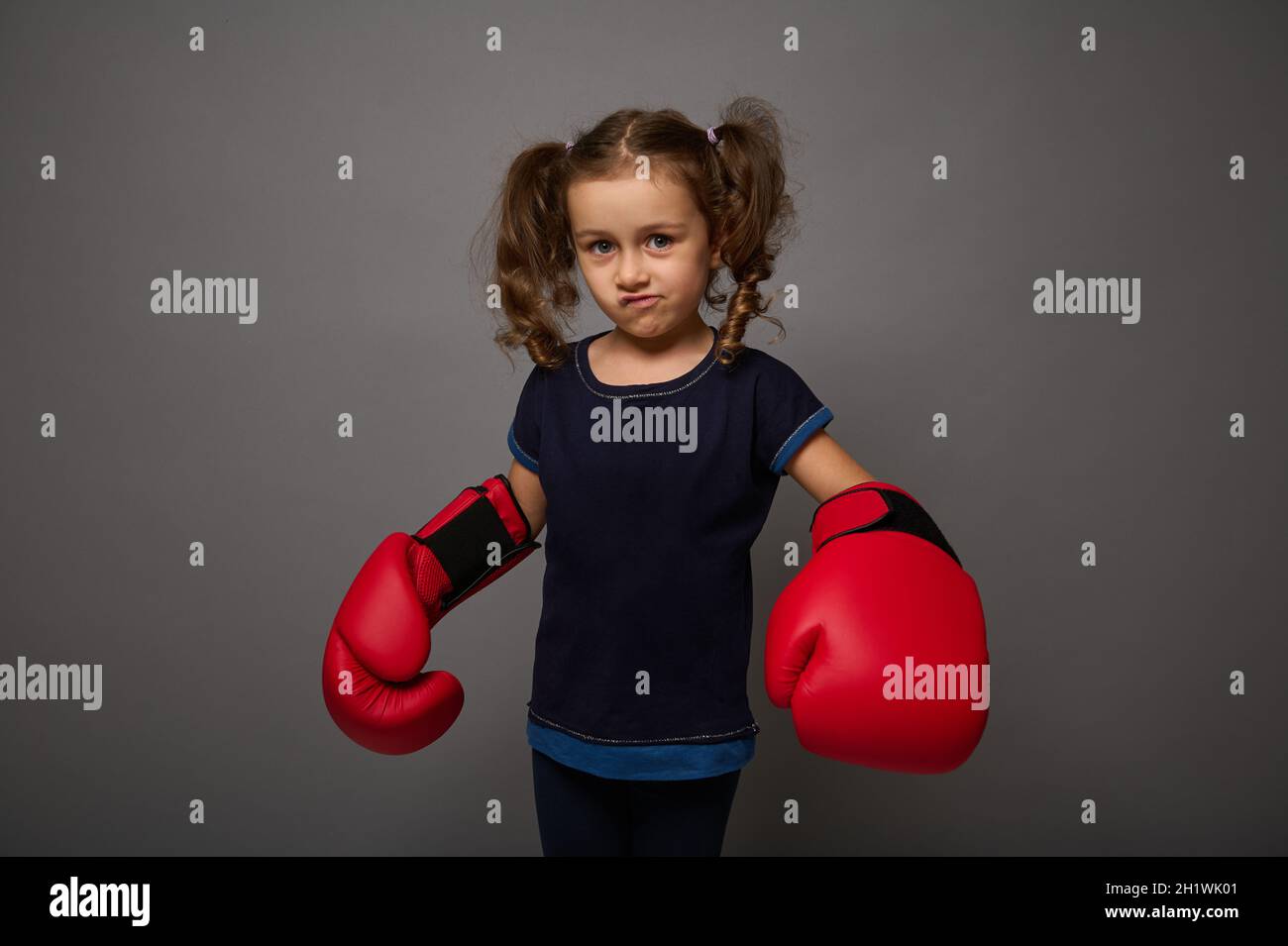 Cute Caucasian baby girl wearing red boxing gloves poses against gray