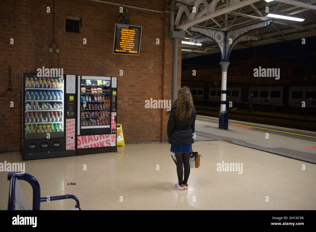 Doncaster, United Kingdom, 22nd May, 2021: Singular young woman wearing a coat, skirt and hand bag, waits during night time at doncaster train station Stock Photo