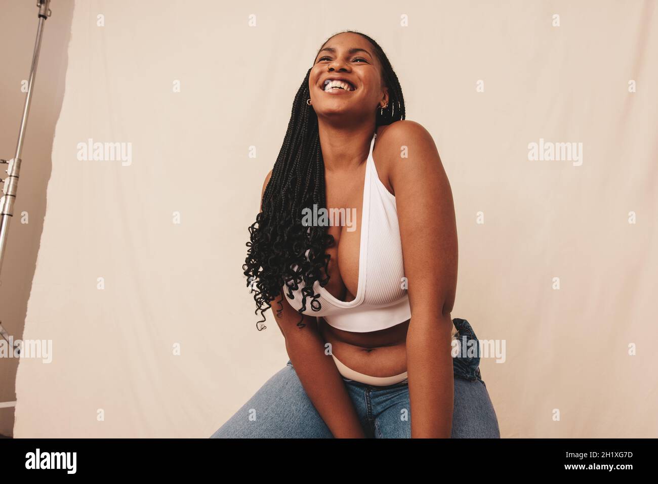 Carefree young woman smiling while sitting on a chair in a studio. Happy young woman feeling comfortable in denim jeans and a bra. Body positive young Stock Photo