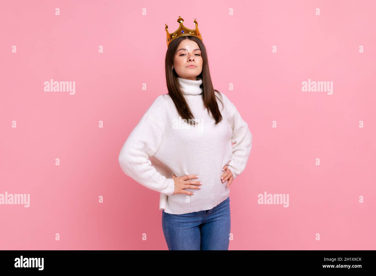 Young female with crown on head, self confidence in success, self-motivation and dreams to be best, wearing white casual style sweater. Indoor studio shot isolated on pink background. Stock Photo