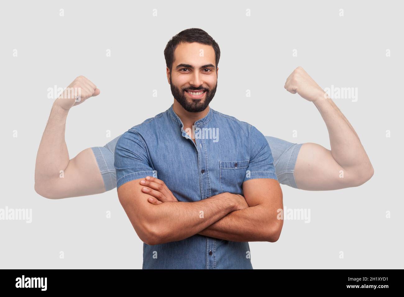 Successful positive bearded man in blue shirt folding hands on chest and looking at camera with toothy smile showing strong big arms in shadow. Indoor studio shot isolated on gray background Stock Photo