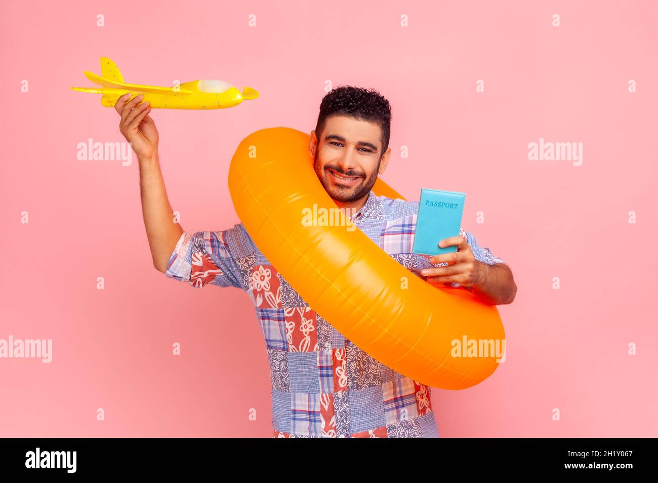 happy satisfied tourist man in blue shirt standing with rubber ring, holding passport document and airplane mockup, enjoying travel tour. Indoor studio shot isolated on pink background. Stock Photo