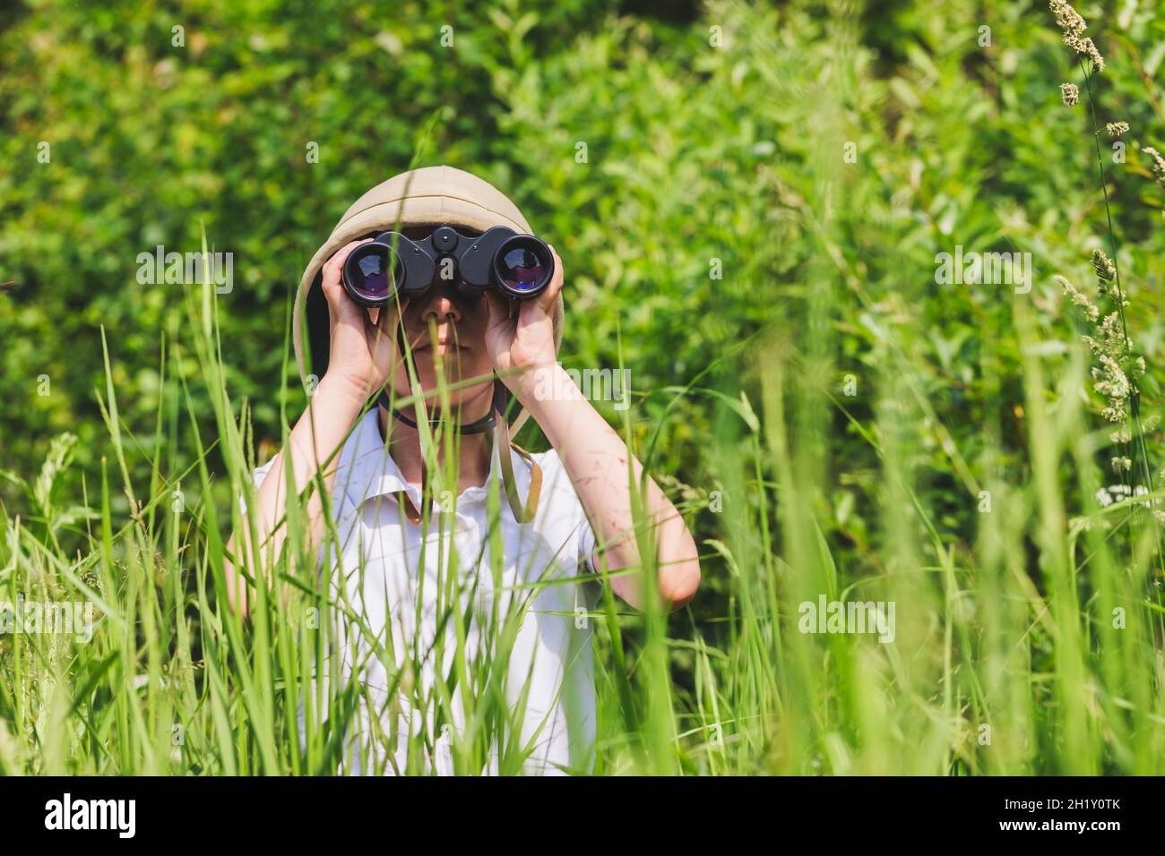 Preteen girl wearing cork helmet standing in grass looking through binoculars. Discovery concept Stock Photo