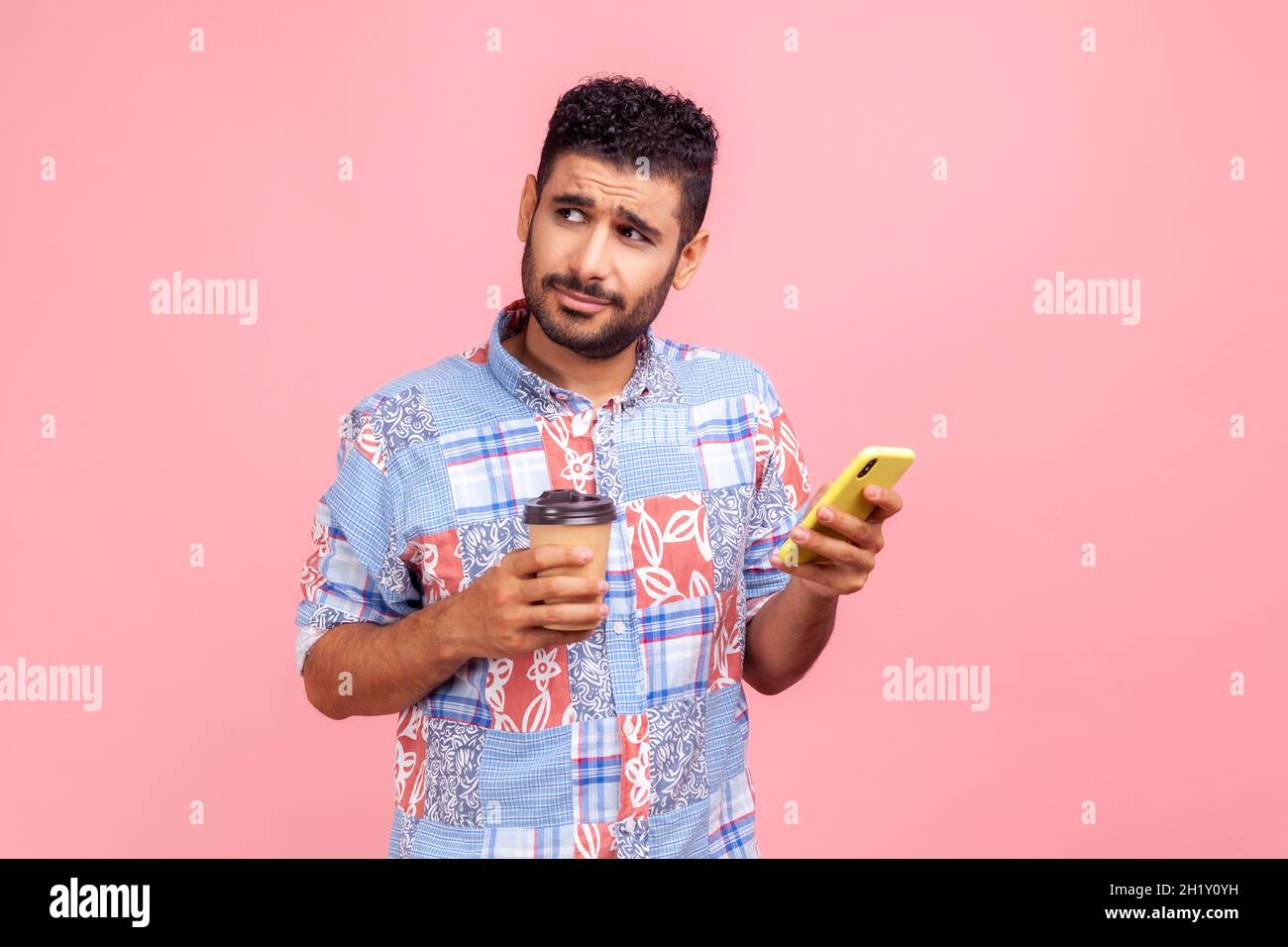 Portrait of handsome pensive young adult blogger man beard in blue shirt standing with coffee and chatting on the phone, looking away. Indoor studio shot isolated on pink background. Stock Photo