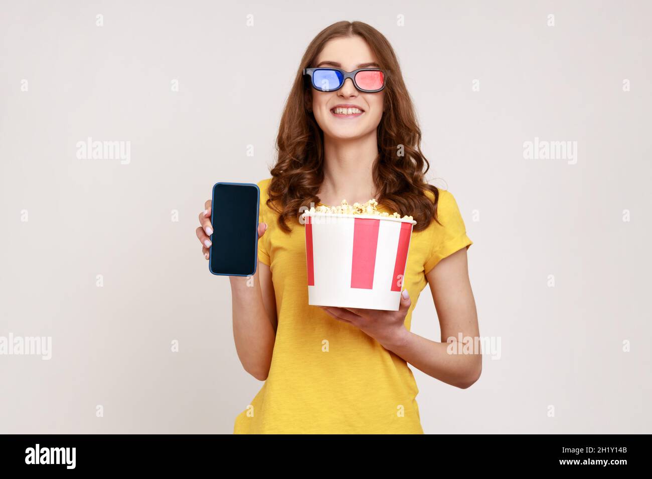 Image of beautiful young woman holding popcorn in bucket, showing smartphone with blank empty screen, smiling to camera, wears yellow T-shirt. Indoor studio shot isolated on gray background. Stock Photo