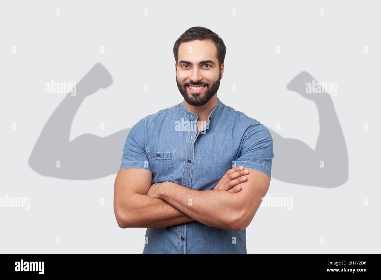 Successful positive bearded man in blue shirt folding hands on chest and looking at camera with toothy smile showing strong big arms in shadow. Indoor studio shot isolated on gray background Stock Photo
