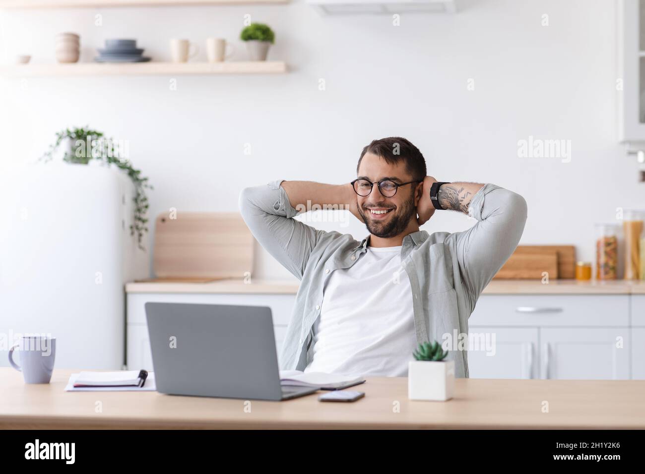 Glad adult caucasian businessman with beard in glasses rest while working at laptop on minimalist kitchen interior Stock Photo