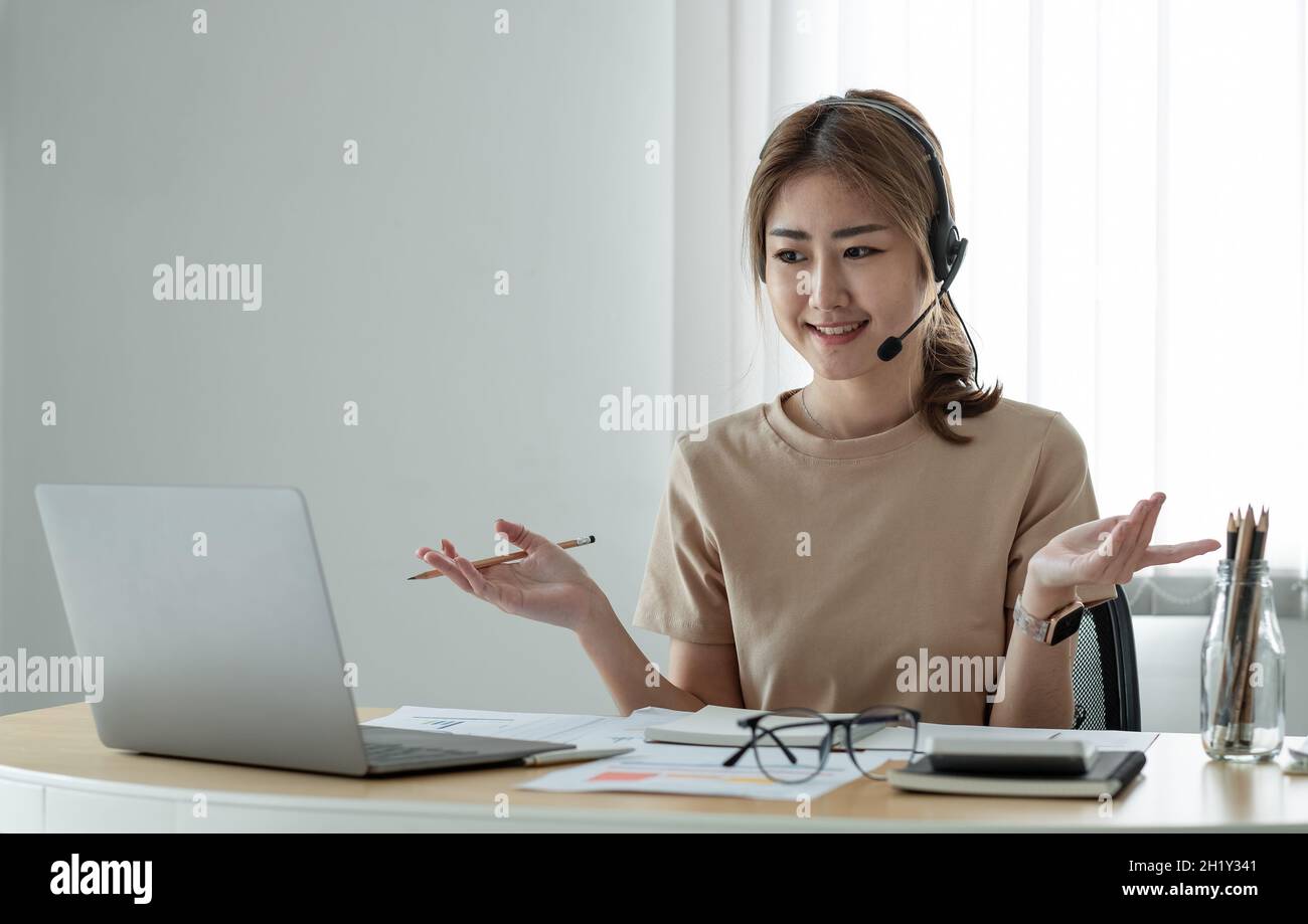 Smiling asian woman freelancer wearing headset, communicating with client via video computer call. Millennial pleasant professional female tutor Stock Photo