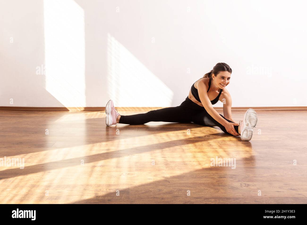 Slim gymnast woman sitting with spread legs, bending to touch feet, doing stretching muscle, wearing black sports top and tights. Full length studio shot illuminated by sunlight from window. Stock Photo