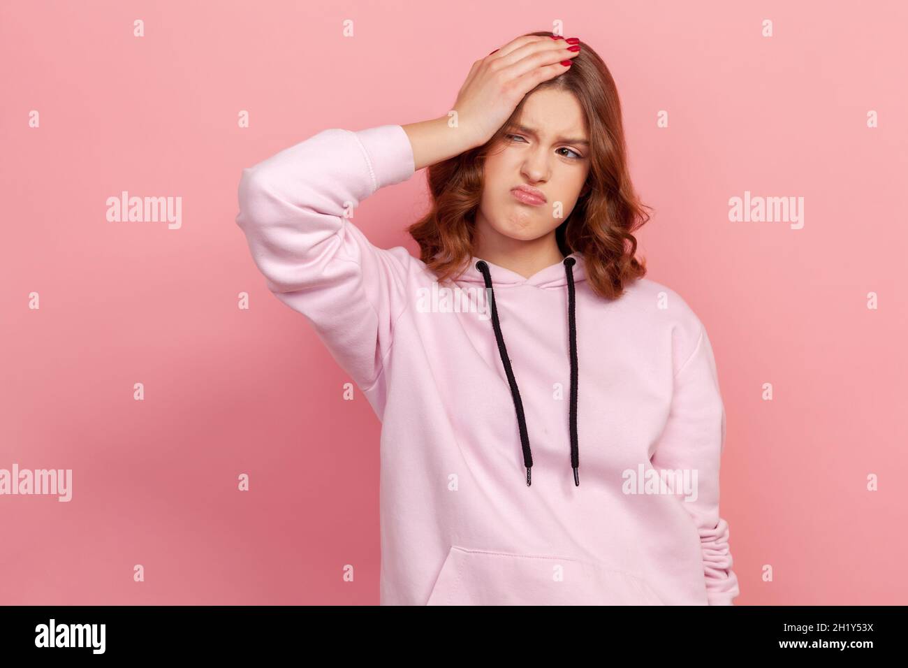 Portrait of young female loser in casual hoodie standing with bad feelings and touching head, worried about problems. Indoor studio shot isolated on pink background Stock Photo