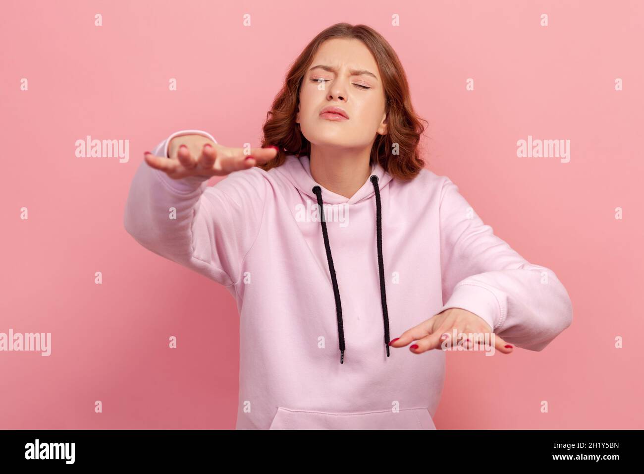 Portrait of concentrated brunette young female in hoodie trying to move with closed eyes and raised hands, vision problems, blindness. Indoor studio shot isolated on pink background Stock Photo