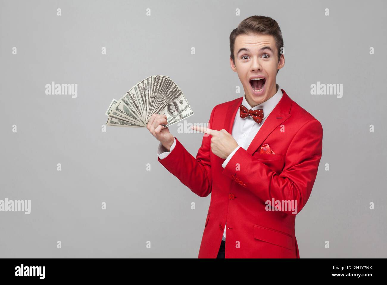 Wow, look at my money. Portrait of surprised man with stylish hairdo in red jacket and bow tie pointing at bunch of dollars, expressing extreme joy and amazement. indoor studio shot, gray background Stock Photo