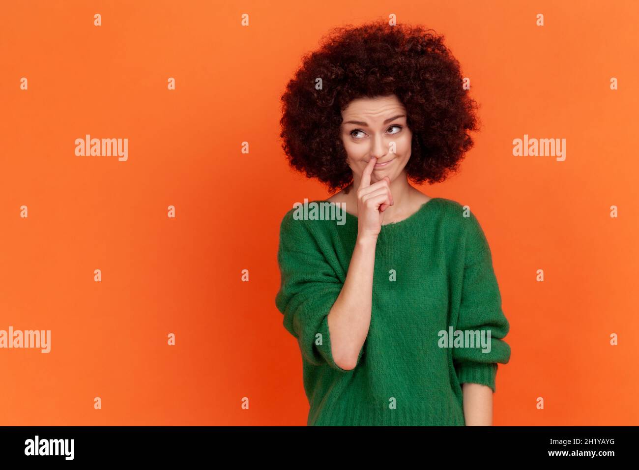Funny woman with Afro hairstyle wearing green casual style sweater picking her nose, demonstrating bad manners, childish behavior. Indoor studio shot isolated on orange background. Stock Photo