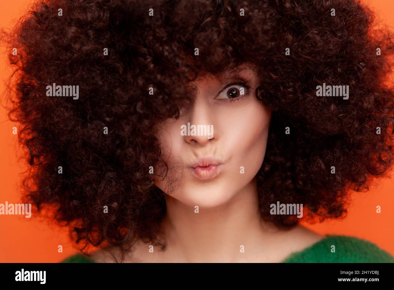 Closeup portrait of woman with Afro hairstyle looking at camera with pout lips, having perfect skin and fluffy hair, advertising beauty salon. Indoor studio shot isolated on orange background. Stock Photo