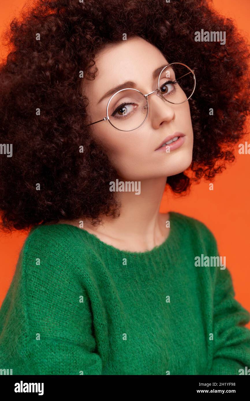 Portrait of pensive woman with perfect skin with Afro hairstyle wearing green casual style sweater, looking at camera, beauty and care concept. Indoor studio shot isolated on orange background. Stock Photo